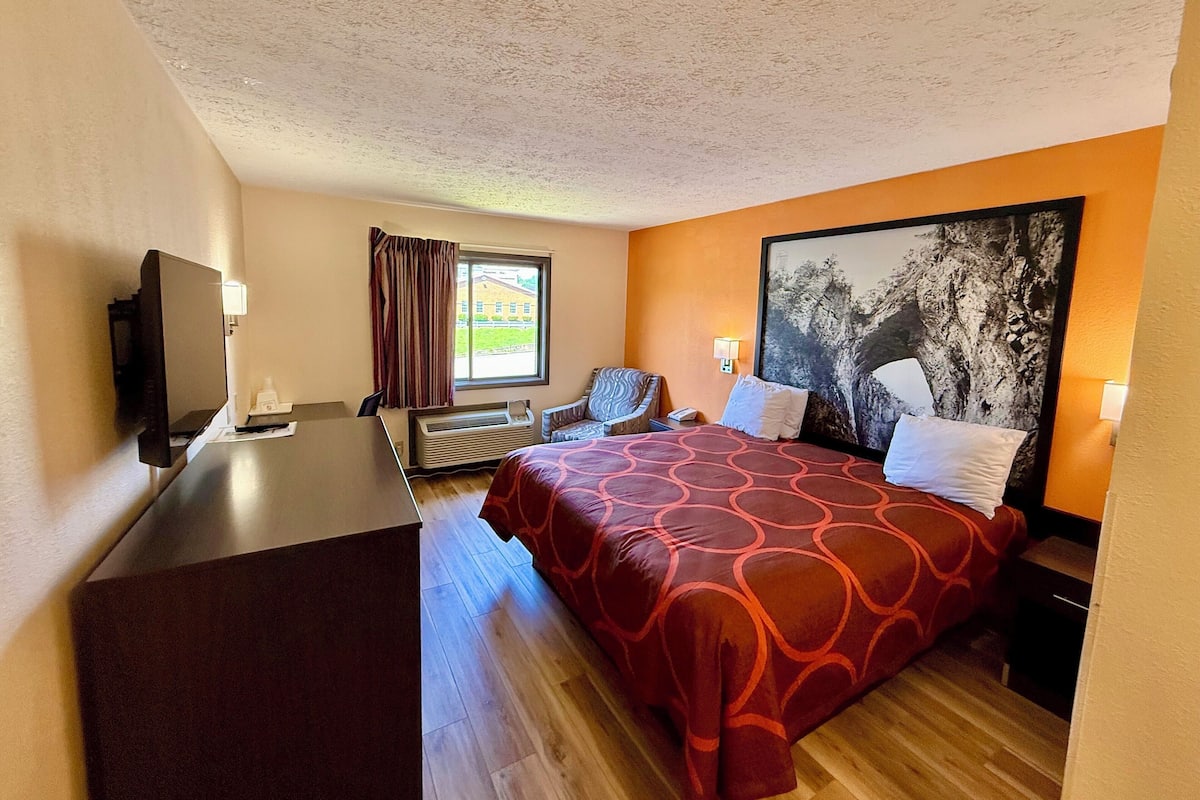 A hotel room with a bed, a desk, a chair, a TV, a window with curtains, and a painting on the wall.