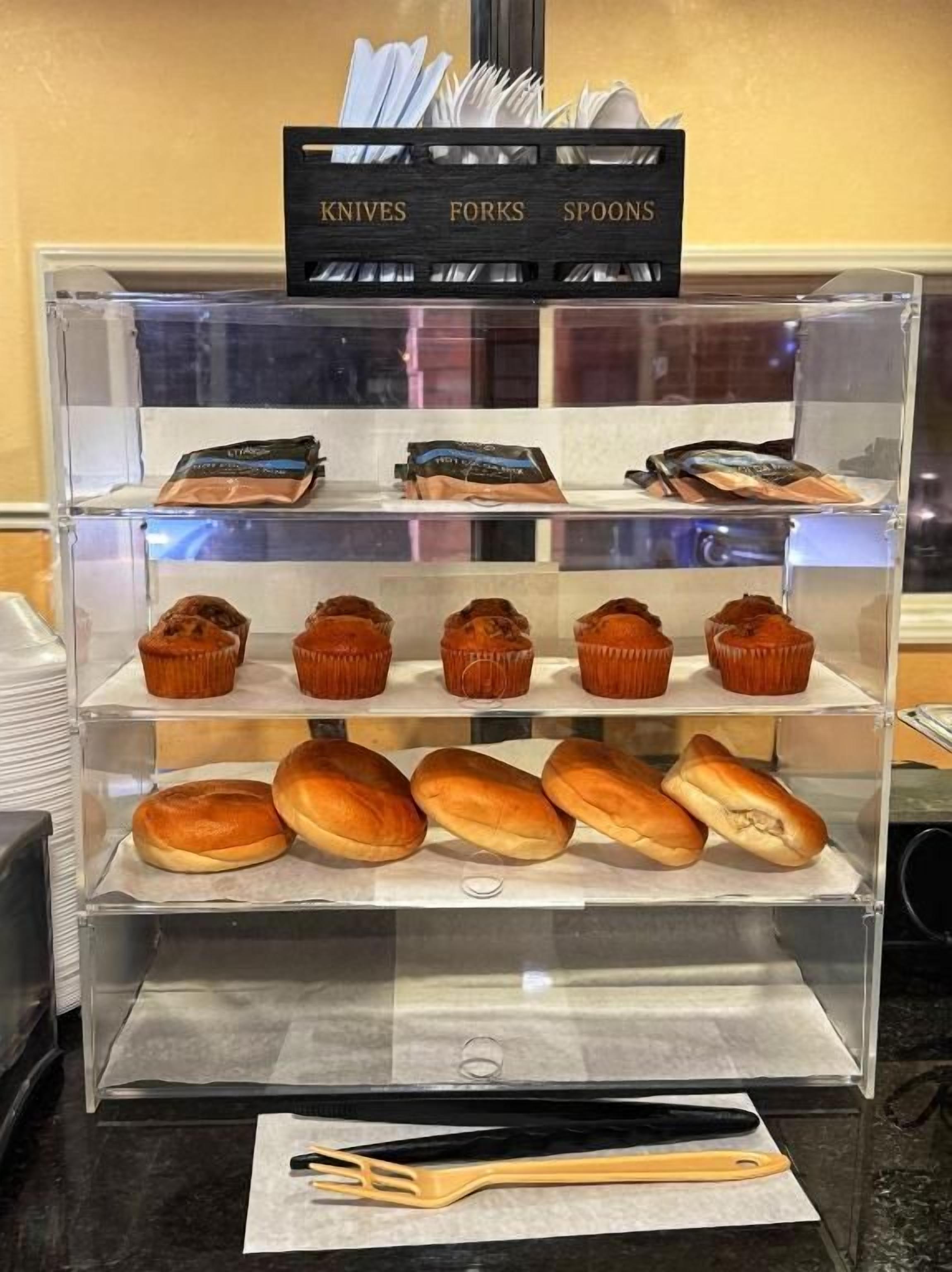 Une vitrine de boulangerie contenant des muffins et des petits pains, et une pancarte au-dessus indiquant où se trouvent les ustensiles.
