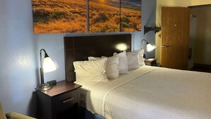 A hotel room with a large bed, two bedside tables with lamps, a wooden door, and a wall-mounted painting of a sunset landscape.