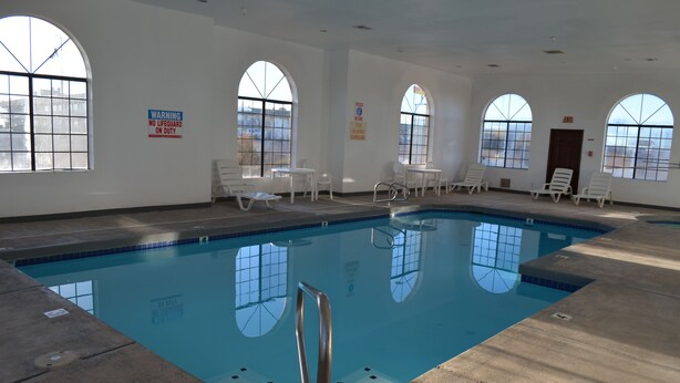 An indoor swimming pool with clear glass windows, white lounge chairs, and a lifeguard warning sign.