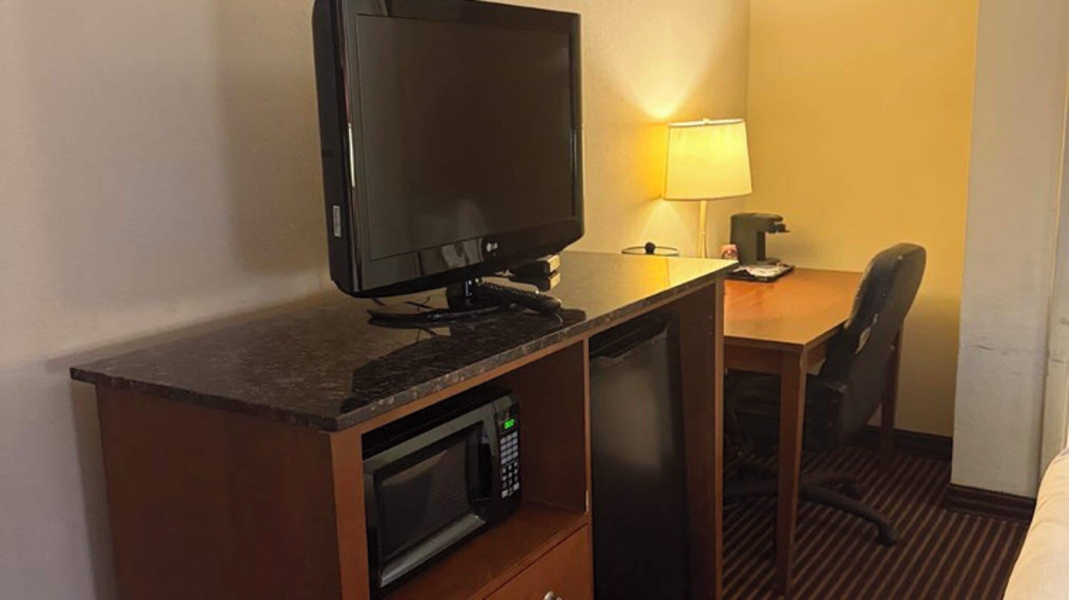 A hotel room with a desk, a television, a microwave, a lamp, and a chair.