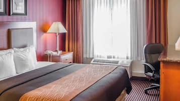 A hotel room with a bed, a desk, a chair, a lamp, and a window with curtains.