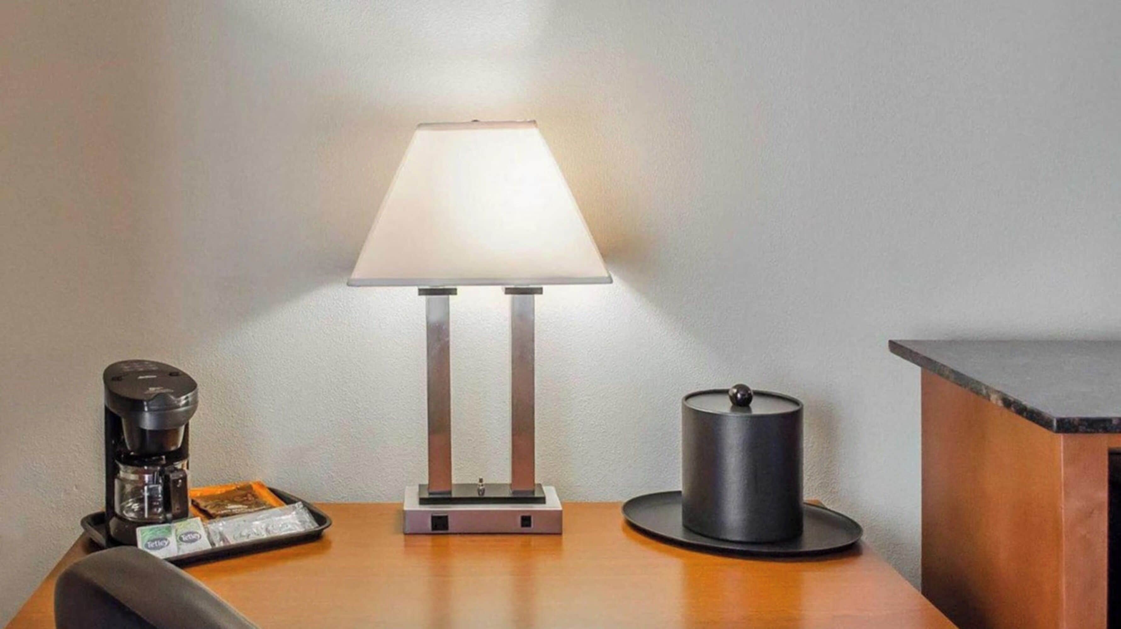 A hotel room with a table featuring a lamp, a coffee maker, and a container.