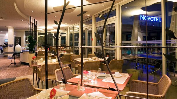 Un restaurante con mesas dispuestas para comer, una barandilla de vidrio y vistas al hotel Novotel.
