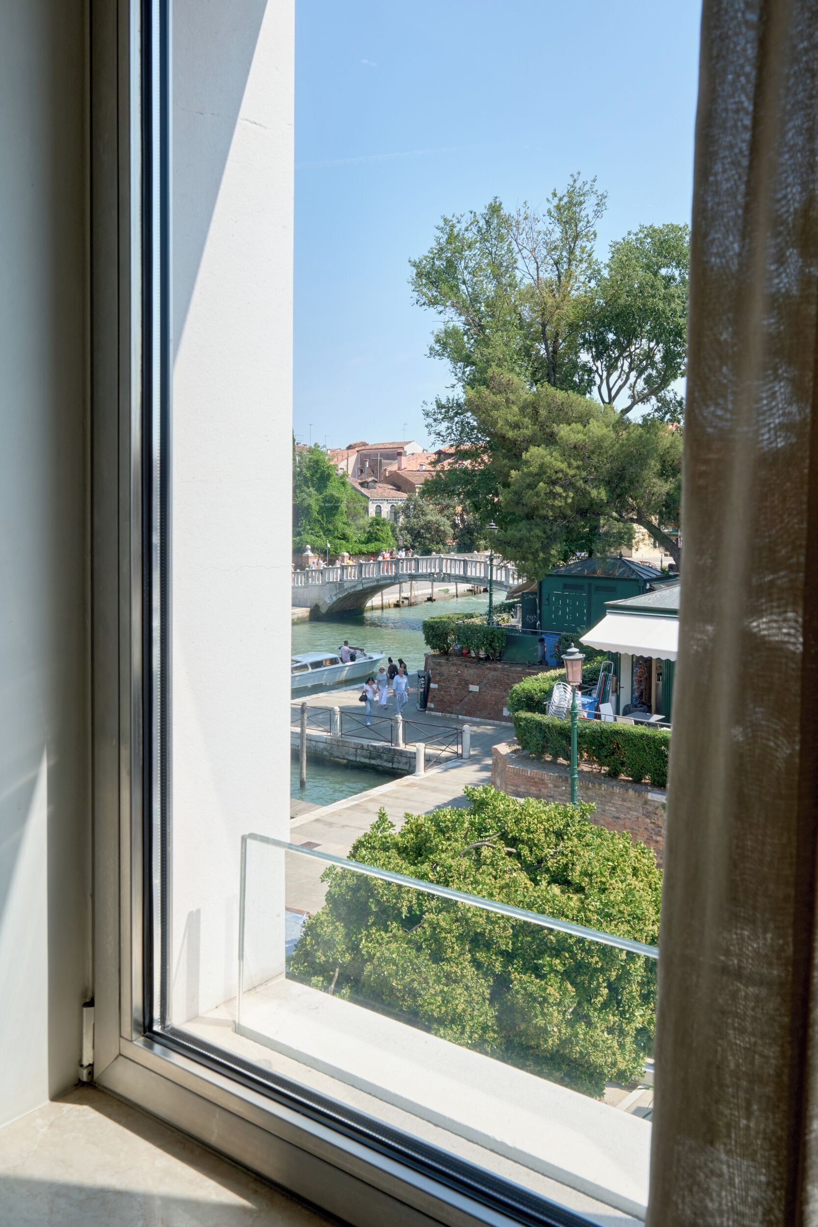 Vista desde una ventana sobre un canal con un puente, vegetación y edificios al fondo.