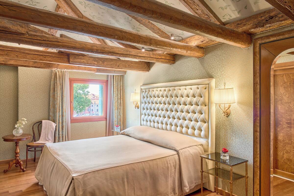 A bedroom with a large bed, wooden beams, a window with curtains, a bedside table with a lamp, and a chair.