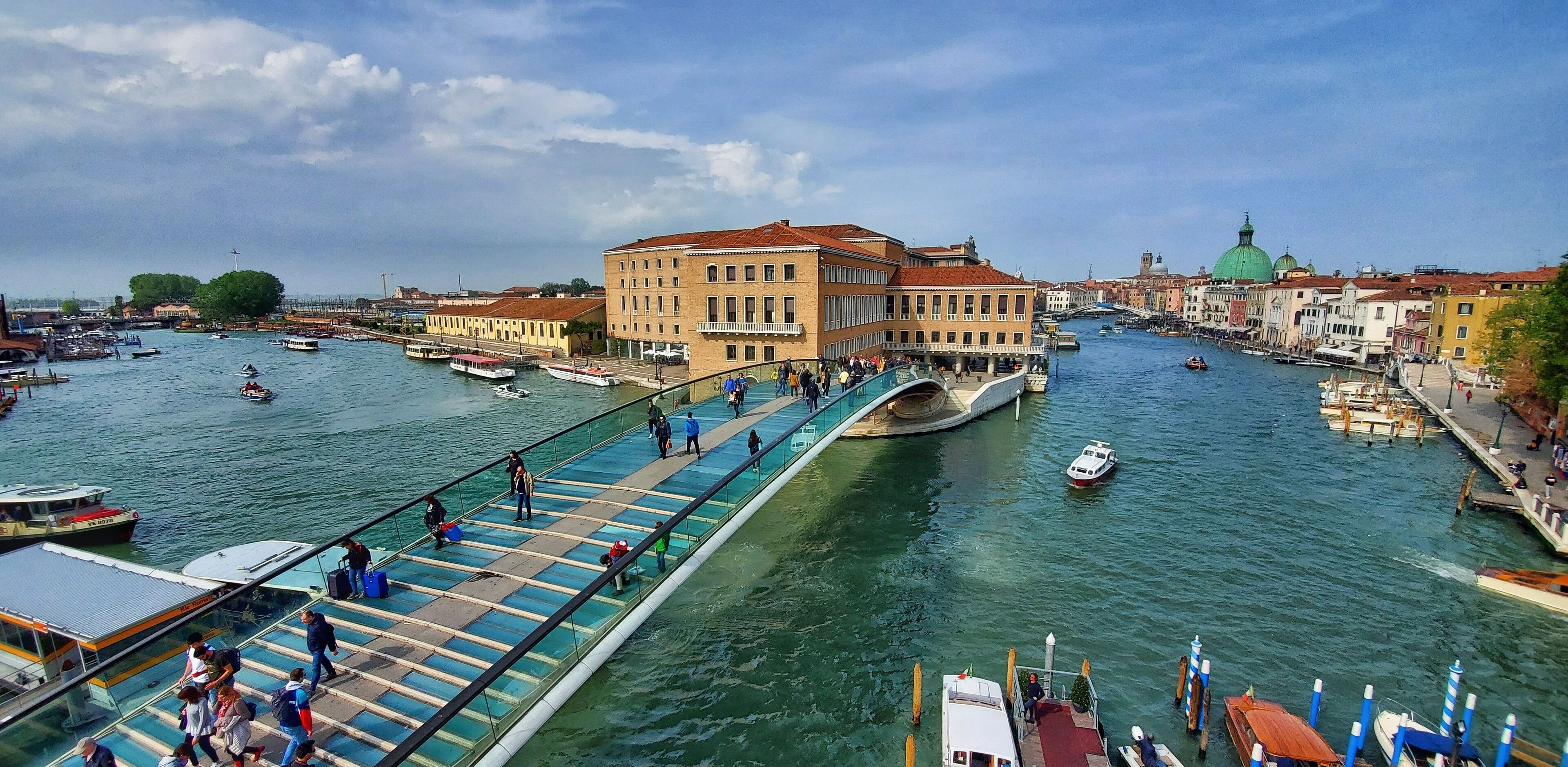 A pedestrian bridge over a river with boats and buildings in the background. - Santa Chiara Hotel (Venice)