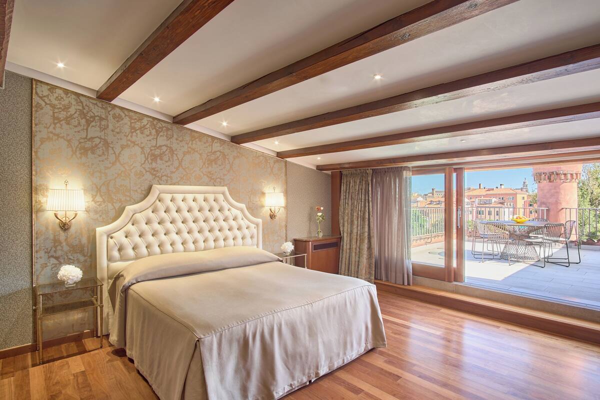 A bedroom with a large bed, wooden beams, a balcony with a table and chairs, and a view of buildings.