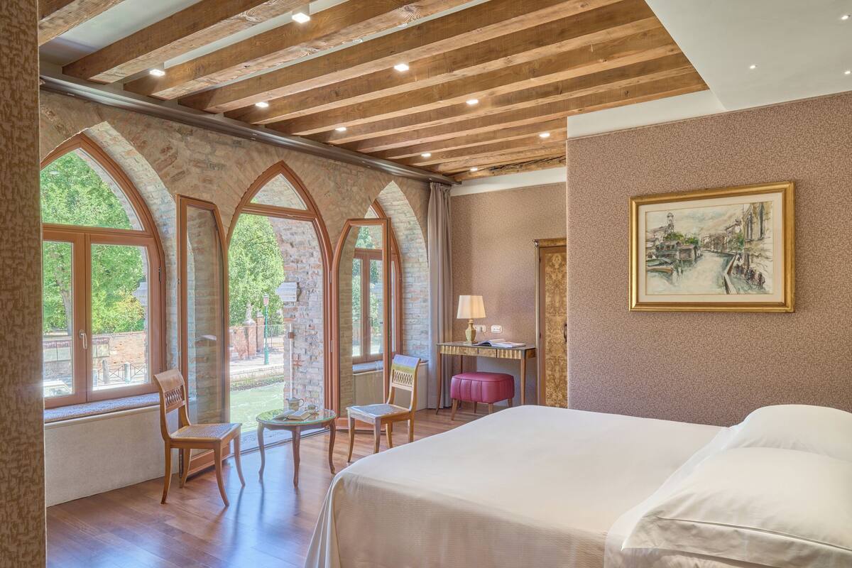 A bedroom with a large bed, wooden beams, stone arches, and a painting on the wall.