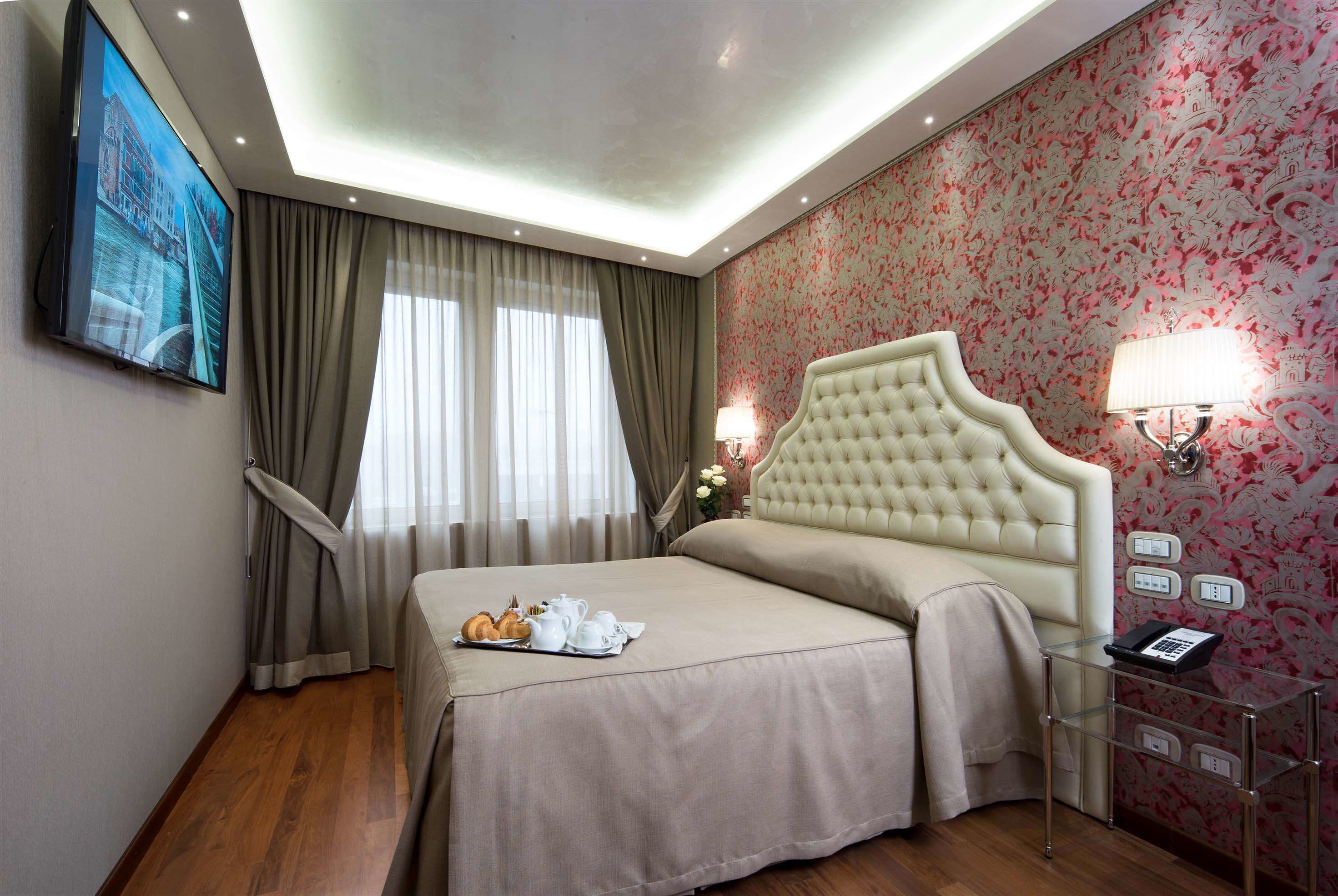 Une chambre d’hôtel dotée d’un grand lit, d’un téléviseur à écran plat fixé au mur, d’une table de chevet avec une lampe et d’un papier peint à motifs floraux.