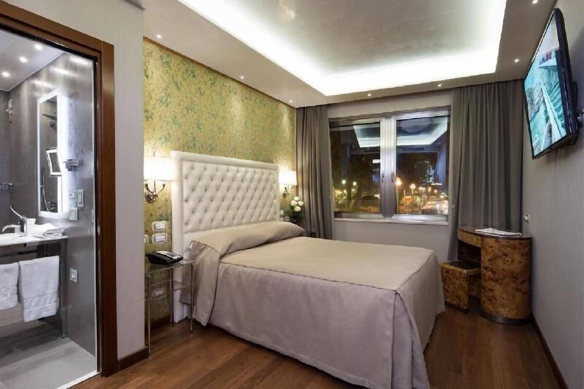 A hotel room with a bed, a vanity with a mirror, a television, and a window with curtains.