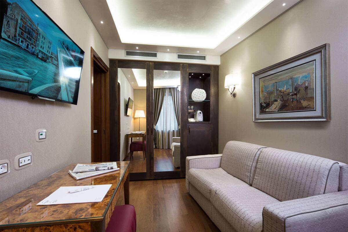 A hotel room with a wooden desk, a cushioned sofa, a television mounted on the wall, and a framed painting.