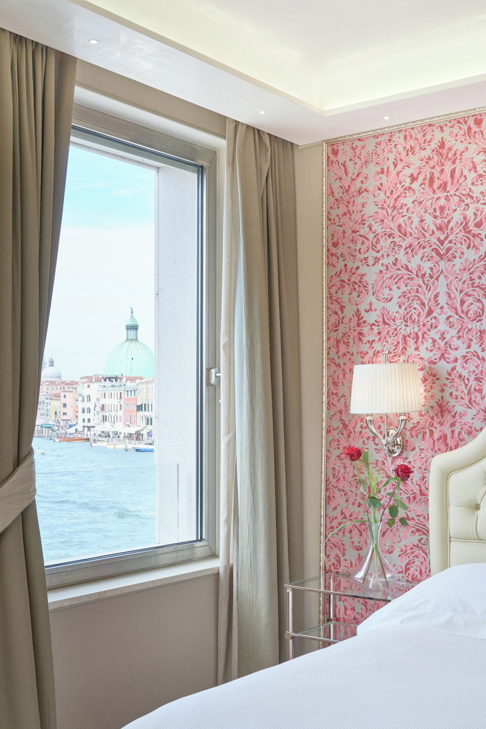 A hotel room with a bed, a nightstand with a lamp, a vase with flowers, and a window with curtains.