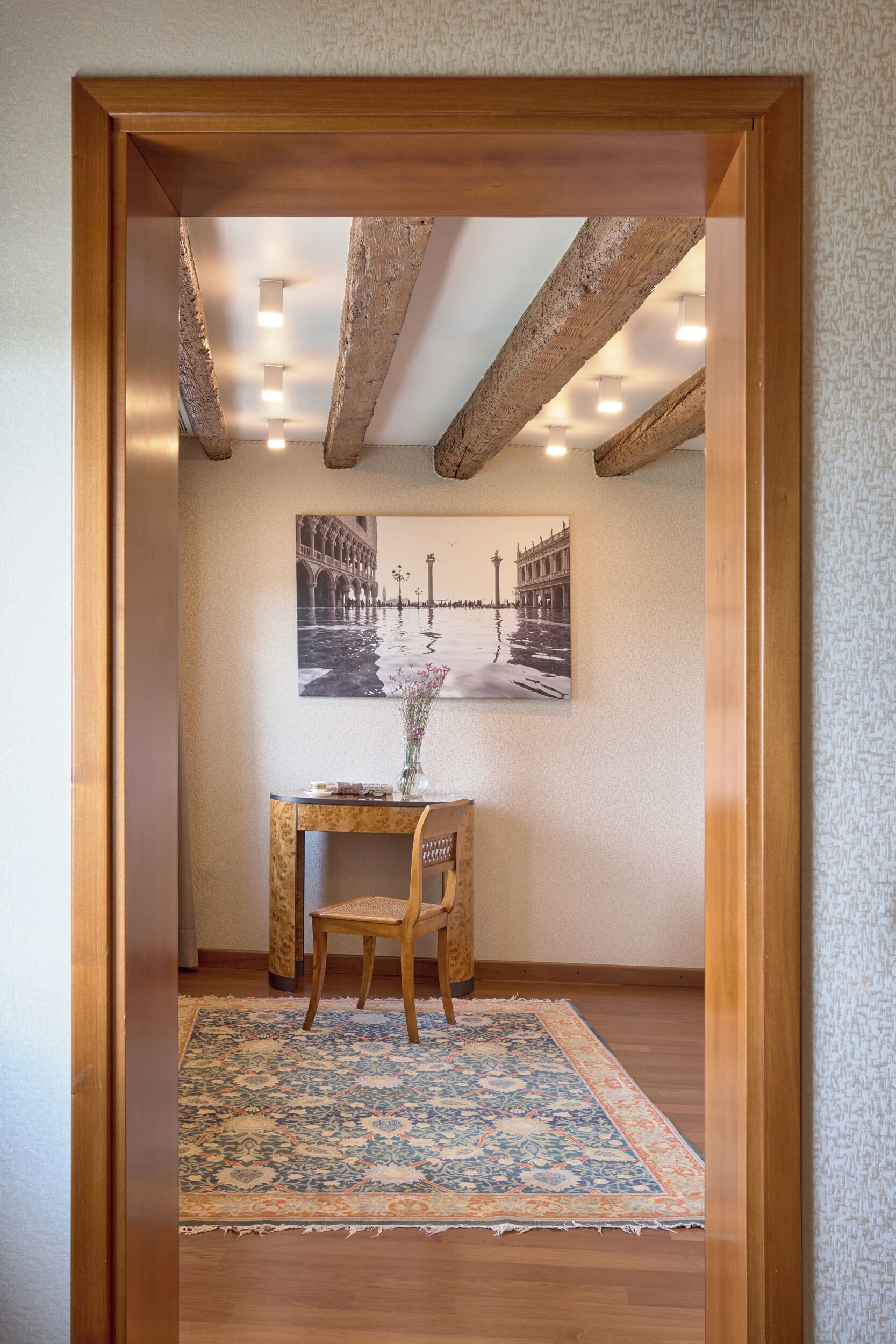 A room with exposed wooden beams, a framed black and white photograph on the wall, a wooden desk with a chair, and a patterned rug on the floor.