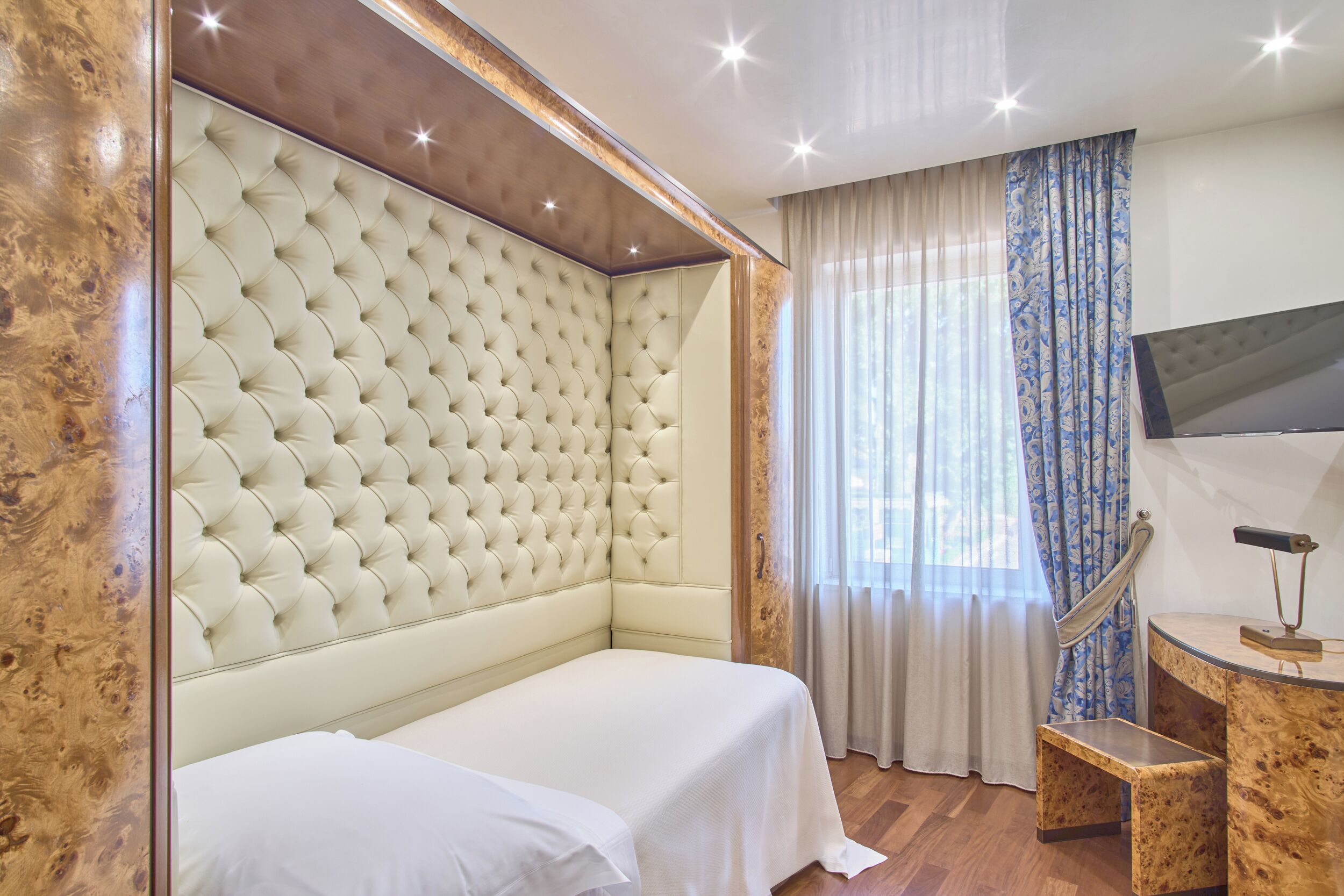 A hotel room with a tufted headboard, a bed with white linens, a wooden side table, a television mounted on the wall, and a window with sheer curtains.