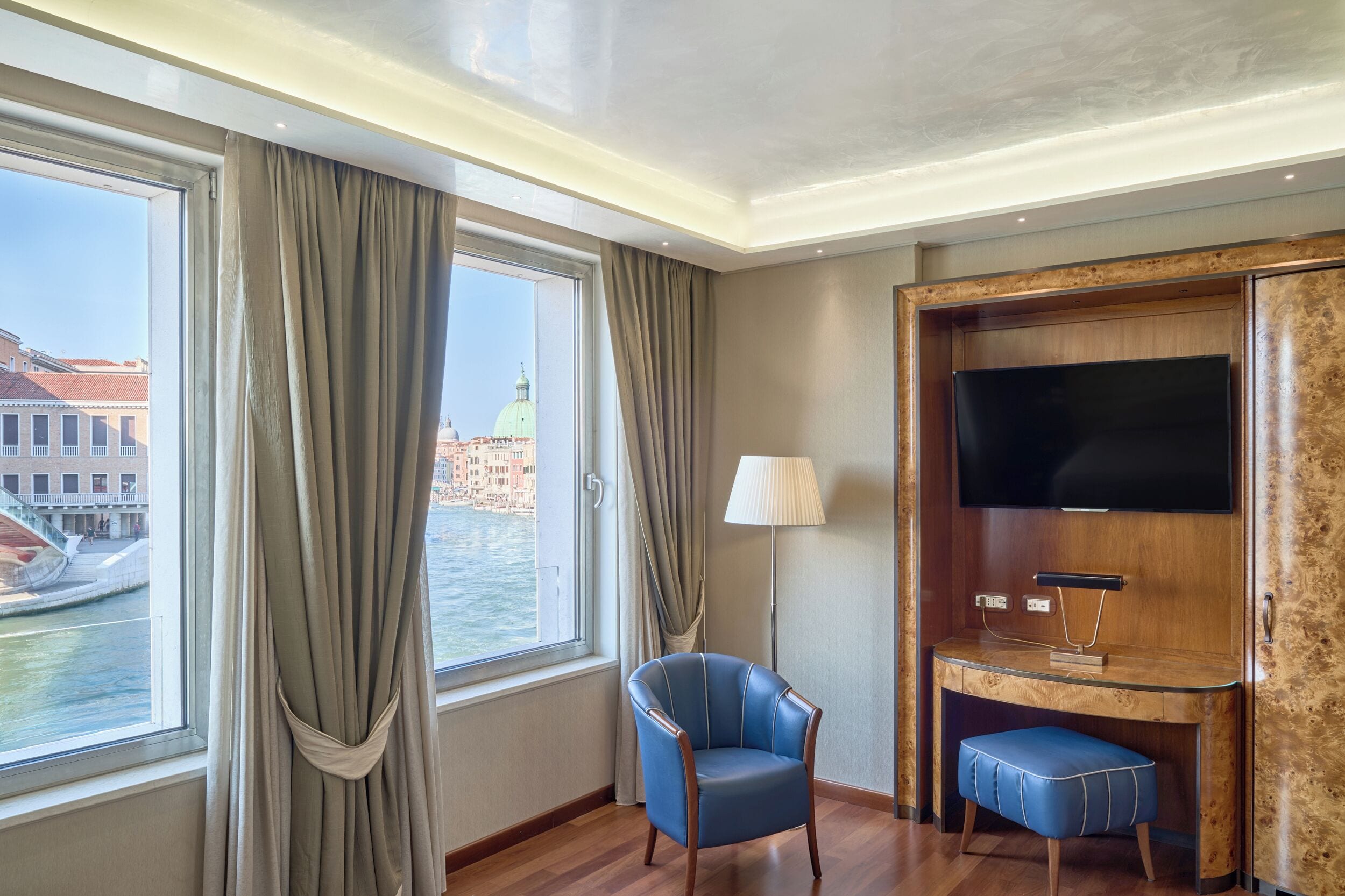 Une chambre d’hôtel avec une grande fenêtre, un fauteuil bleu, un bureau avec un téléviseur à écran plat et une lampe.