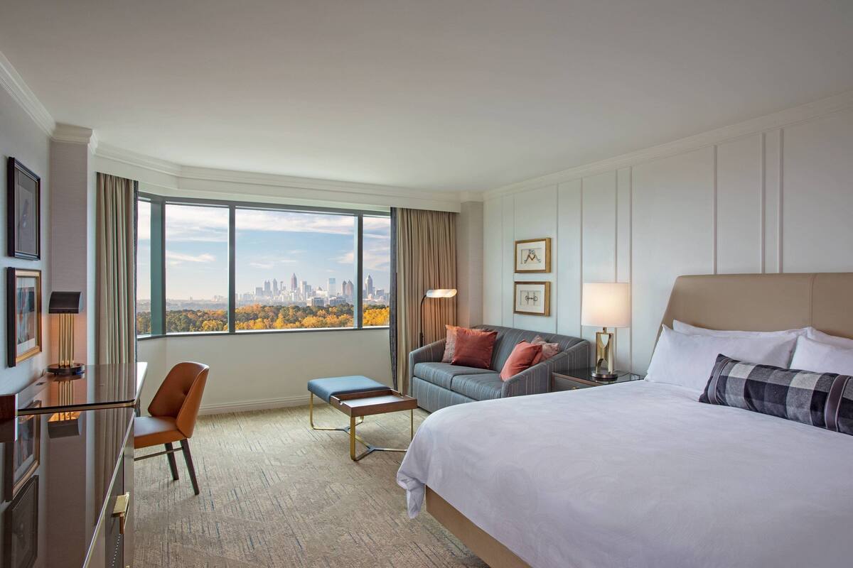 A hotel room with a large bed, a desk, a chair, and a view of a cityscape through large windows.