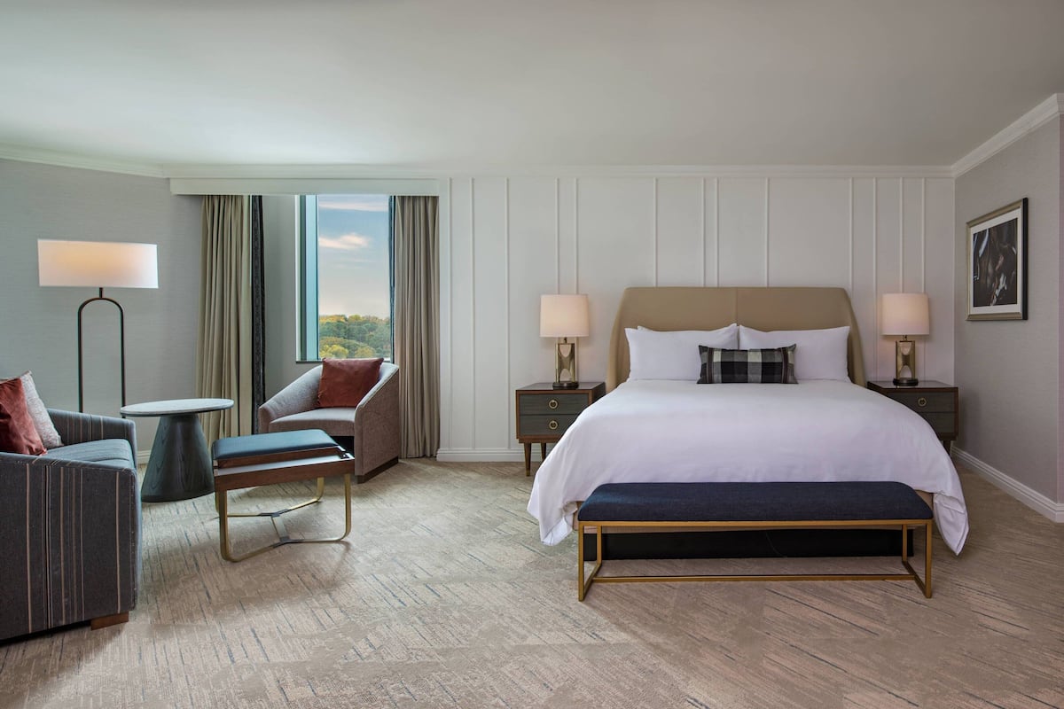 A hotel room with a large bed, two bedside tables with lamps, a sofa, a chair, and a view of the outdoors.
