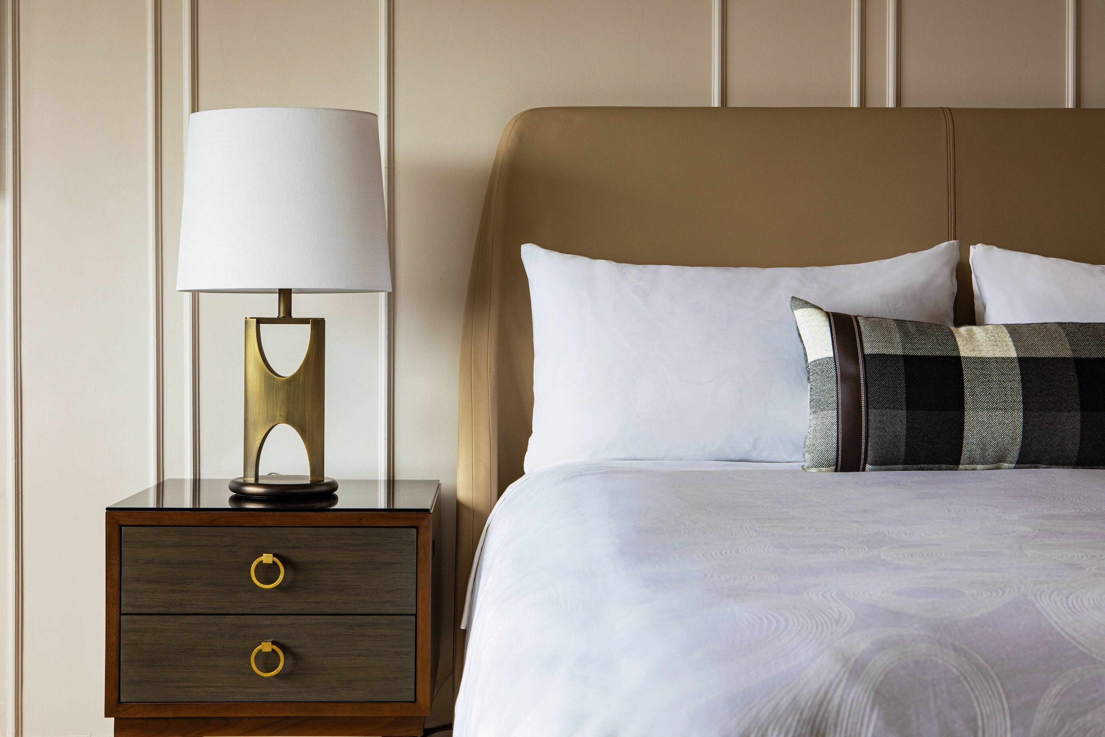 Een netjes opgemaakt bed met witte lakens en een geruit kussen, een bedlamp met gouden voet, en een nachtkastje van hout met twee lades.