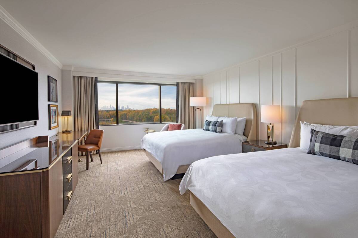 A hotel room with two beds, a desk with a TV, a chair, and a large window offering a city view.