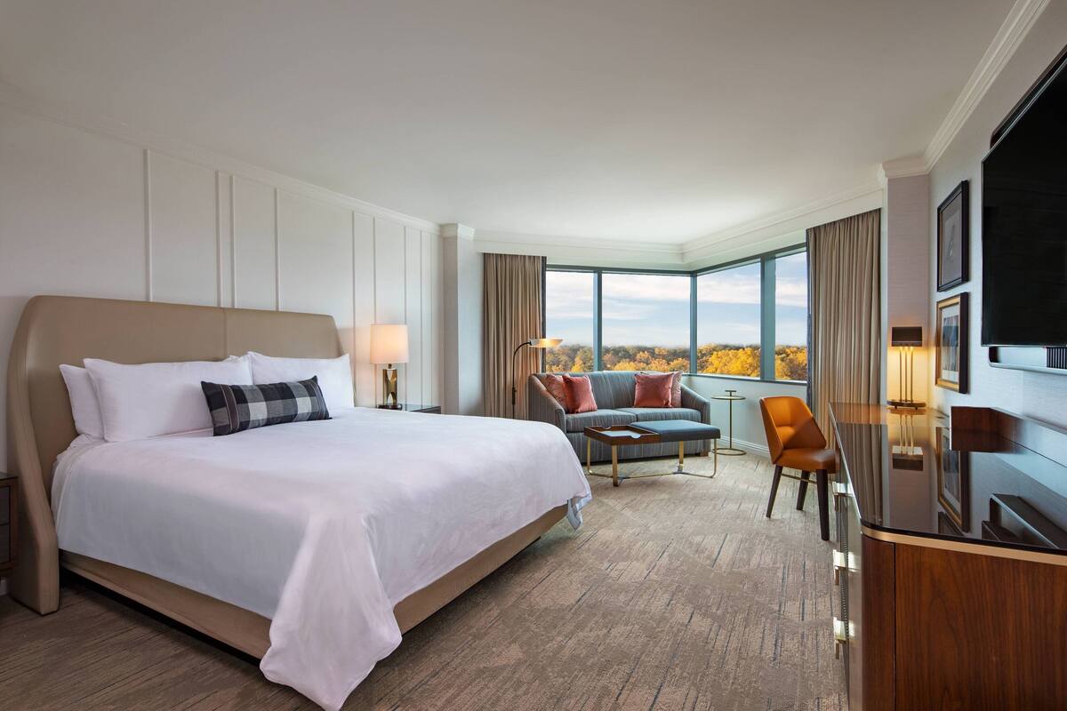 A hotel room with a large bed, a sofa, a TV, and a view of autumn foliage through large windows.