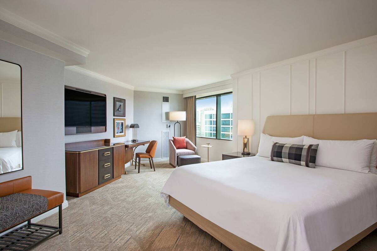 A hotel room with a large bed, a desk with a chair, a sofa, a TV, and a window with a view of buildings.