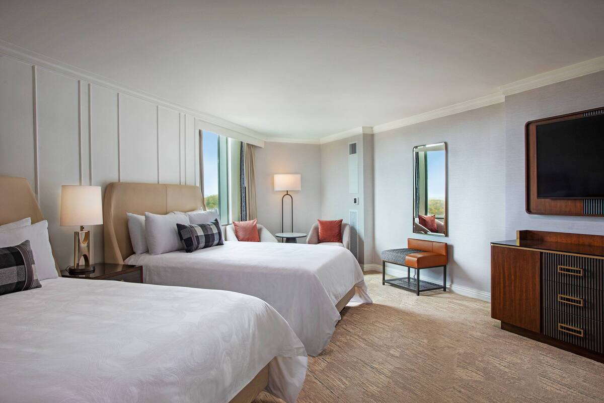 A hotel room with two beds, a desk, a chair, a TV, and a large mirror.