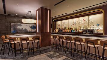 A bar area with a round bar, multiple stools, a large illuminated display, and a brick wall with a painting.
