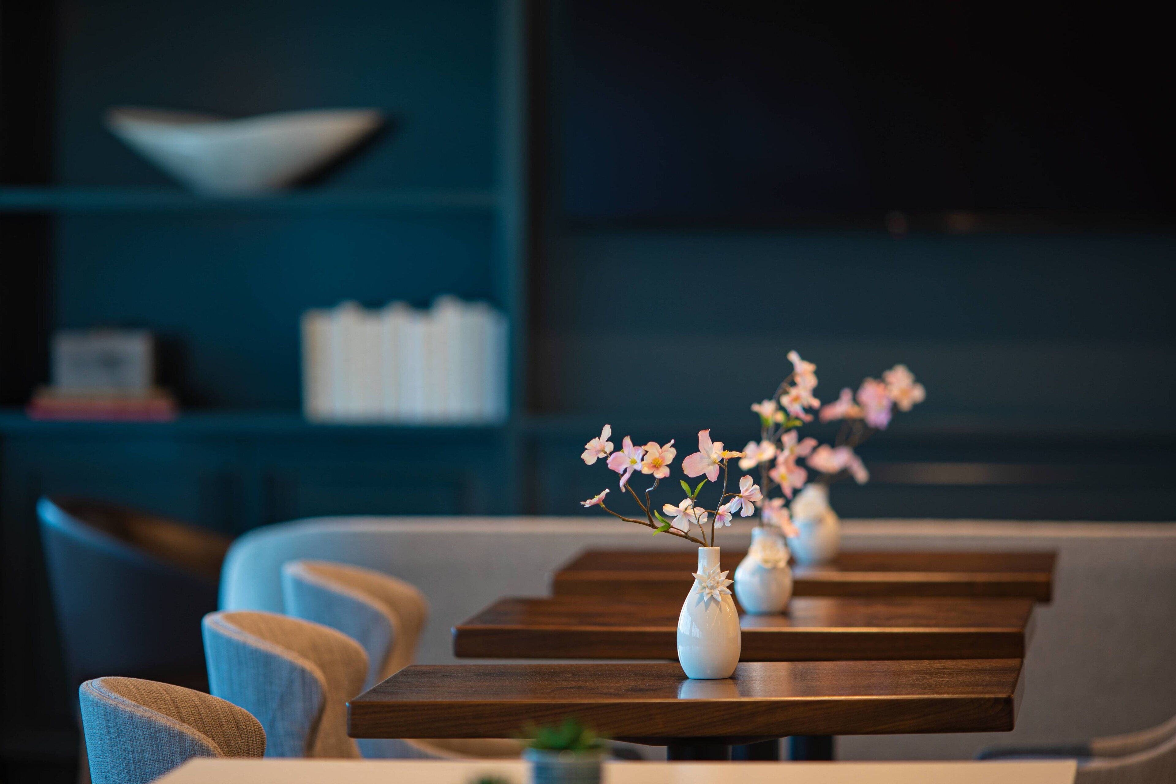 A row of wooden tables with white vases containing pink flowers, set against a blue wall with a white shelf in the background.