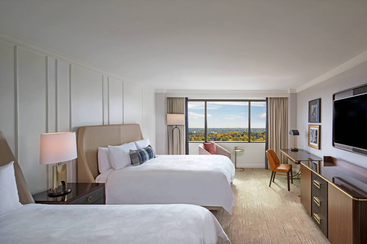 A hotel room with two beds, a desk, a TV, and a window with a city view.
