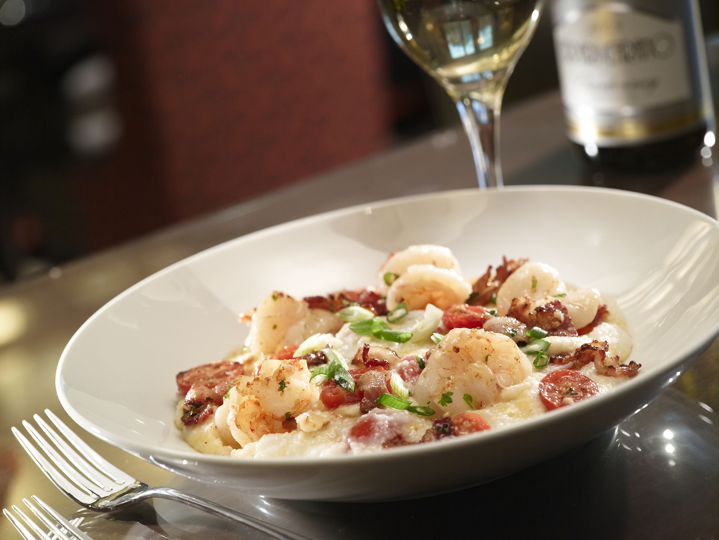 A plate of food with shrimp and bacon, a glass of white wine, and a bottle of wine in the background.