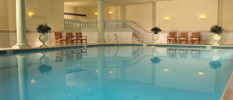 An indoor swimming pool with clear blue water, surrounded by white columns and chairs, and a glass ceiling above.