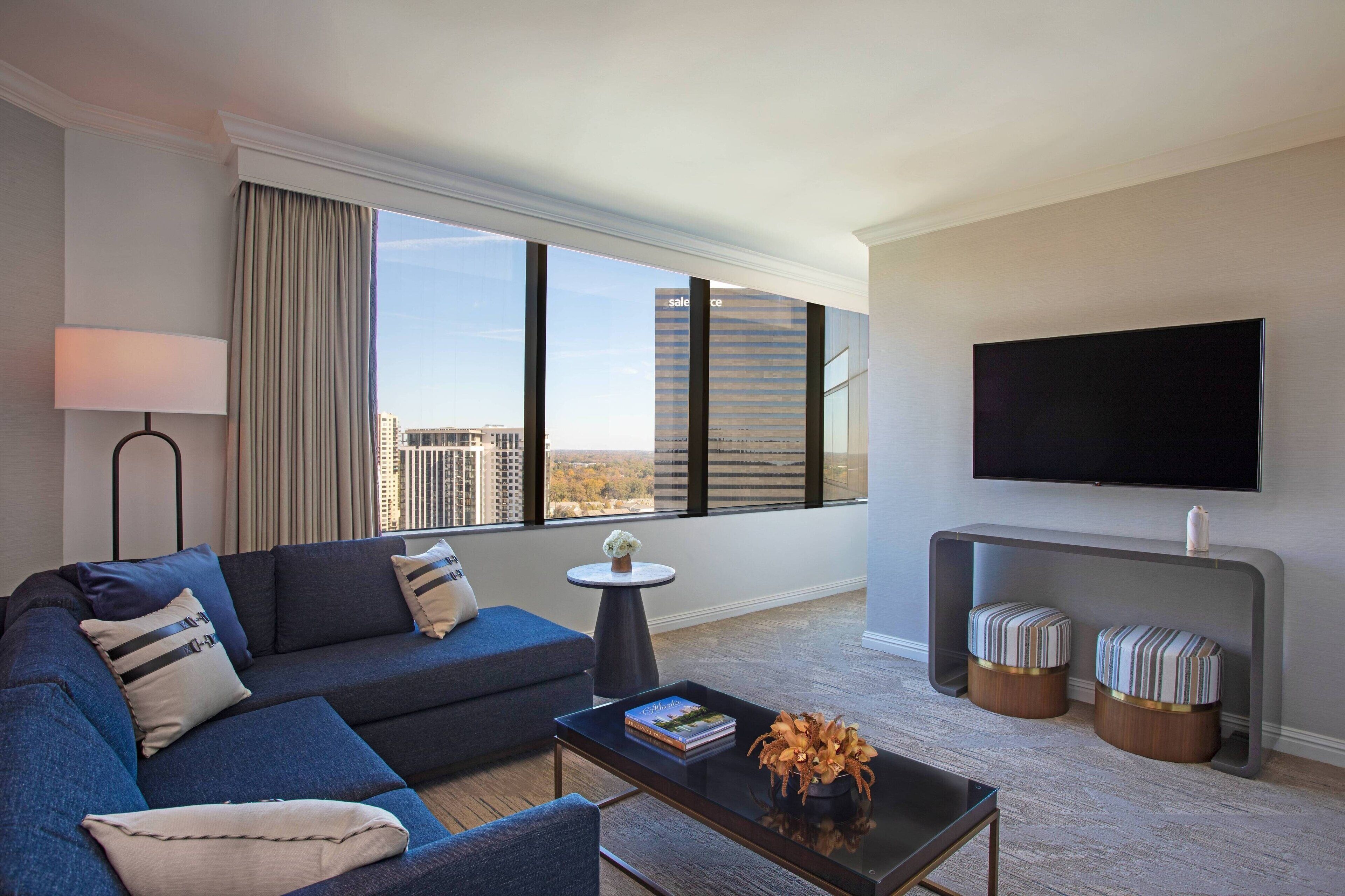 A modern hotel room with a large window, a flat-screen TV, a sofa, a coffee table, and two ottomans.