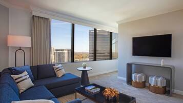 A modern hotel room with a large window, a flat-screen TV, a sofa, a coffee table, and two ottomans.