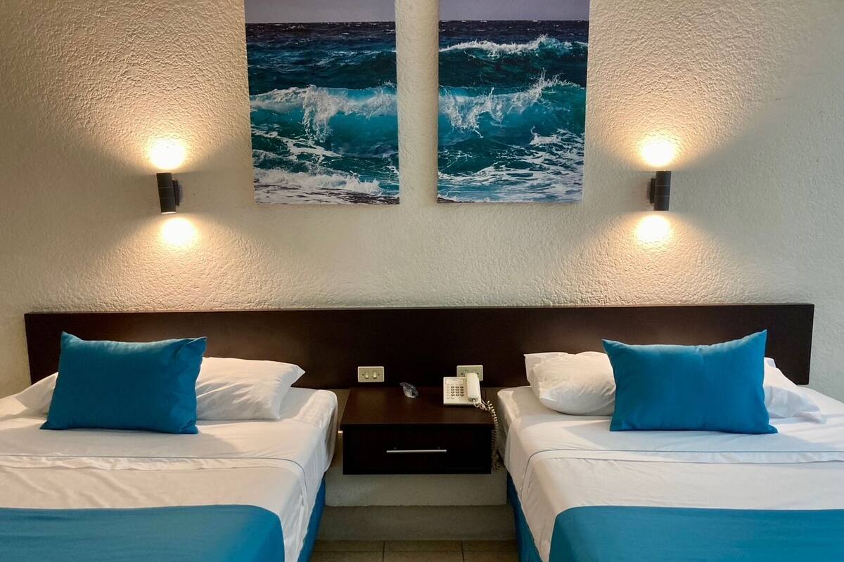 Ein Hotelzimmer mit zwei Betten, jeweils mit blauer Bettdecke und Kissen, einem Nachttisch mit Telefon und zwei Wandleuchten.