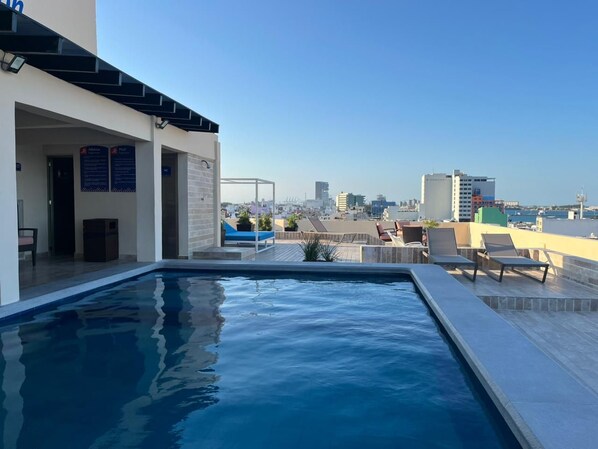 Una piscina en la azotea con sillones de descanso, vistas despejadas del horizonte urbano y un edificio de diseño moderno.