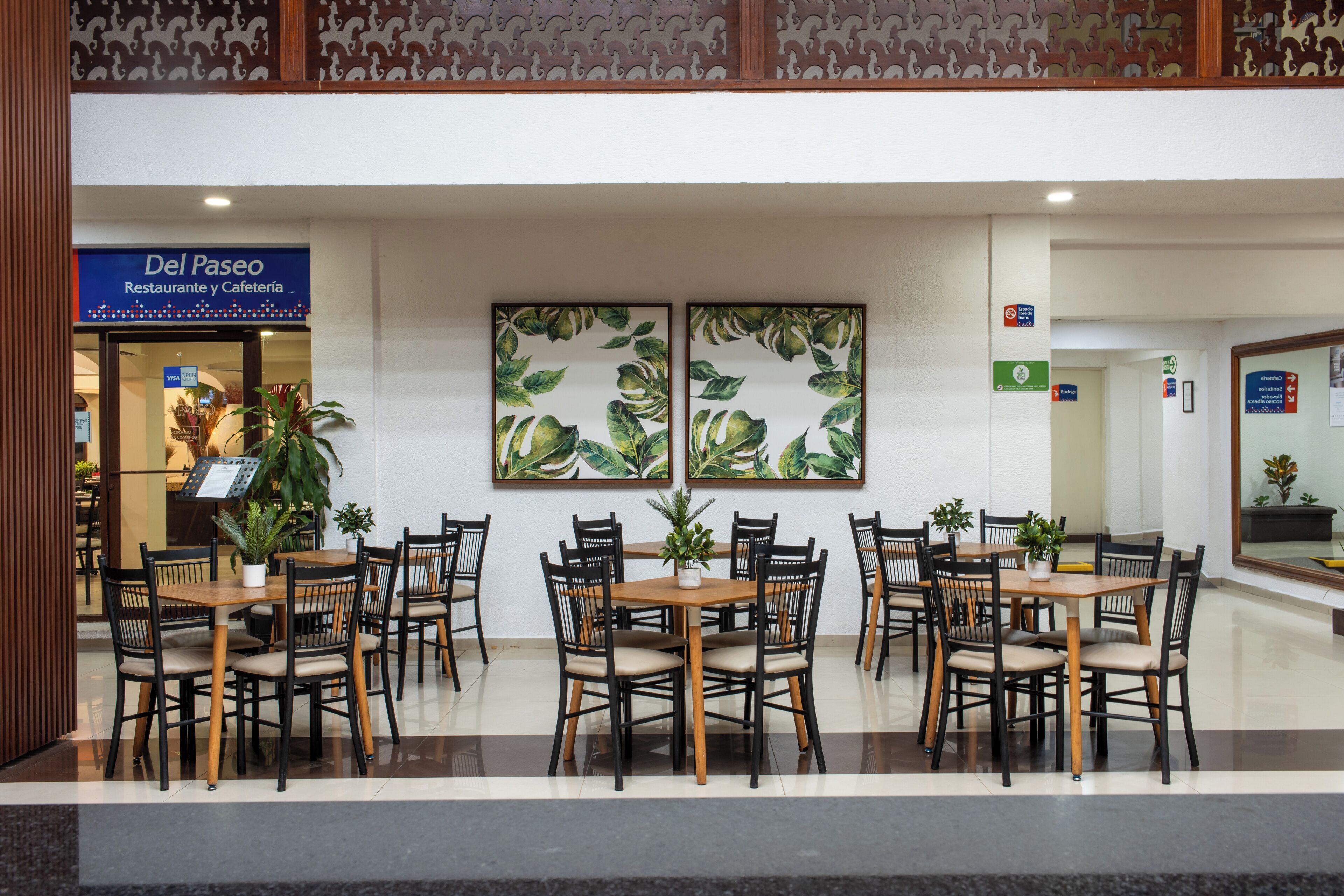A restaurant with multiple tables and chairs, a sign for 'Del Paseo Restaurante y Cafeteria', and decorative wall art.