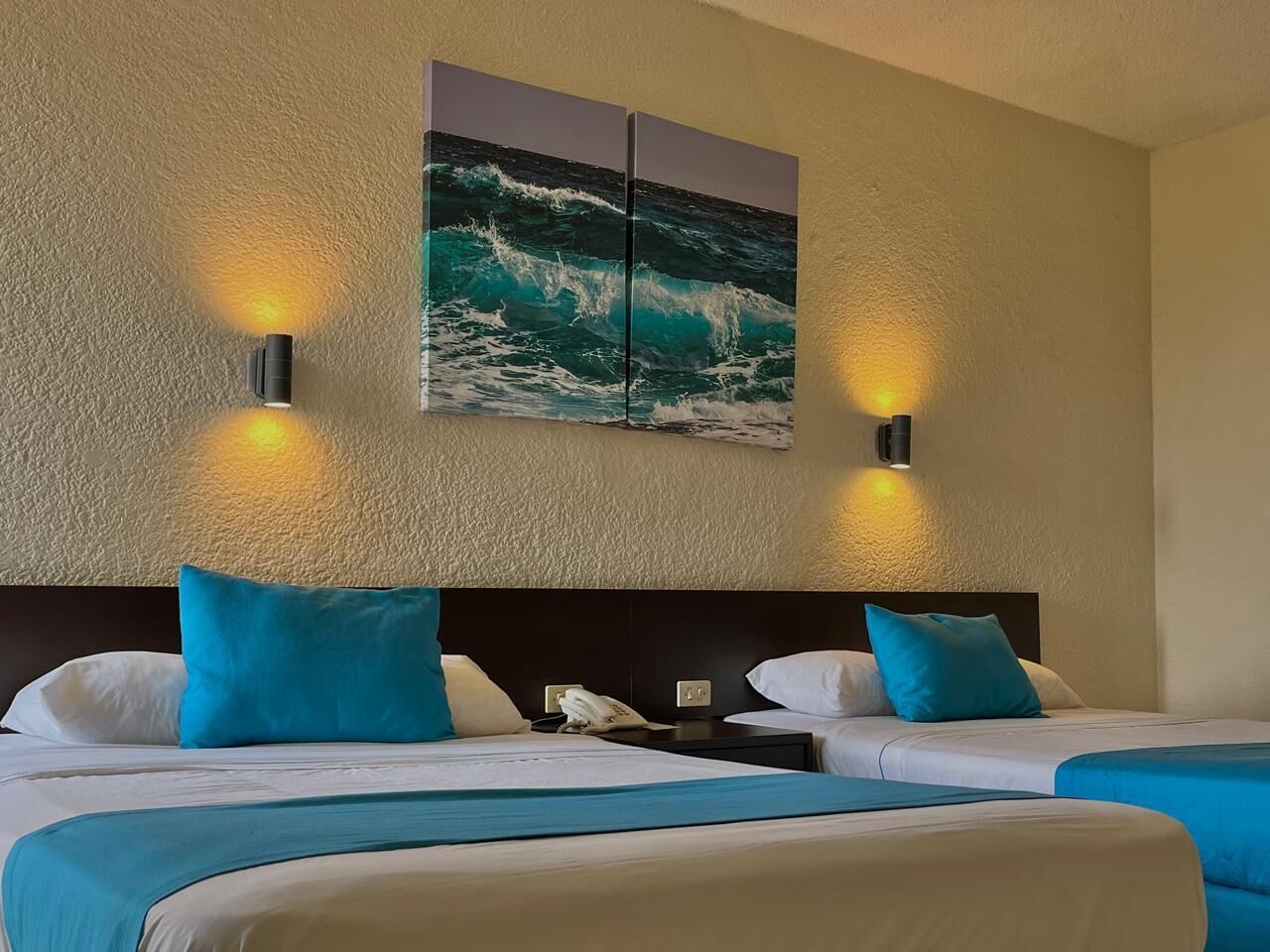Habitación de hotel con dos camas, un cuadro grande de olas en la pared y dos luces fijadas a la pared.