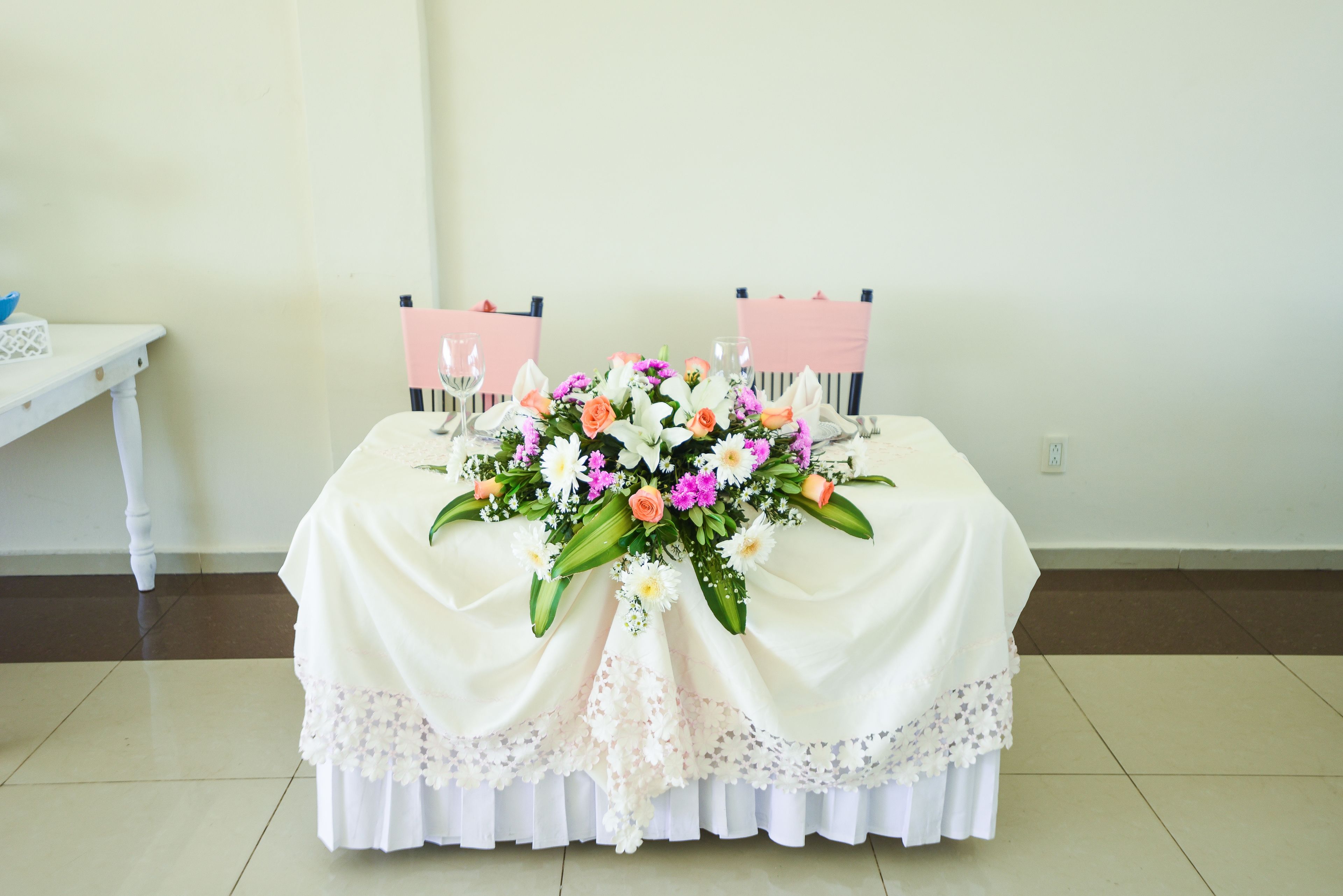 Una mesa con mantel blanco, un centro de mesa floral y dos sillas rosas.