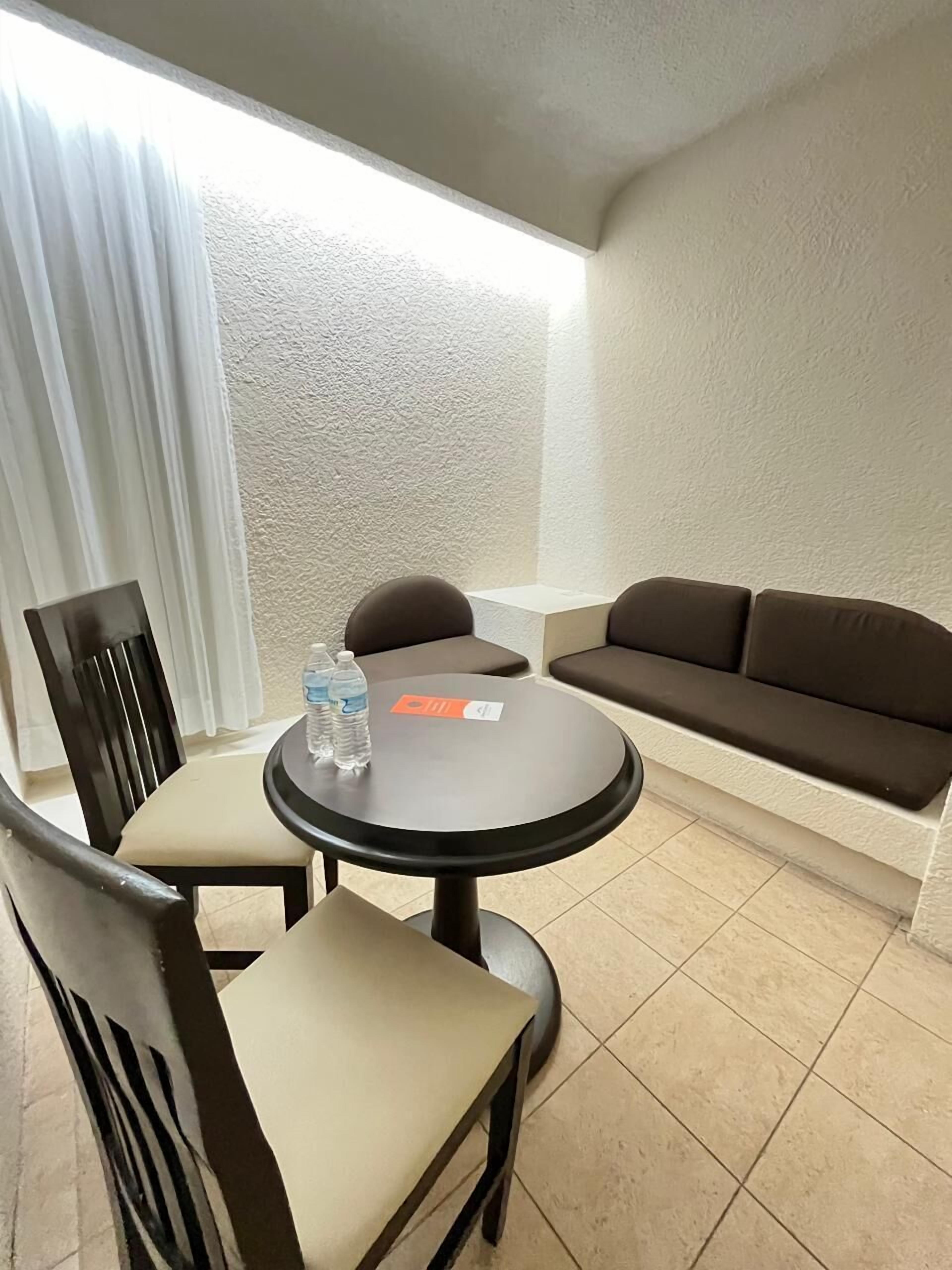 A small, furnished room with a round table, two chairs, a sofa, and a window with curtains.