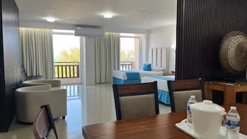 Habitación de hotel con una mesa de comedor, dos sofás, una cama con ropa de cama azul y un balcón con cortinas.