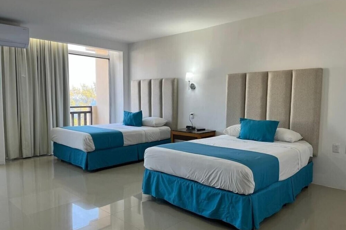 Habitación de hotel con dos camas, una mesita de noche, una lámpara y vistas al exterior.