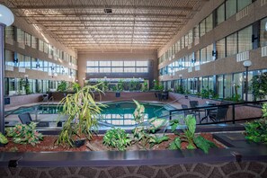 Amplia zona de piscina interior con un techo que presenta un diseño geométrico, rodeada de plantas y zonas de descanso.