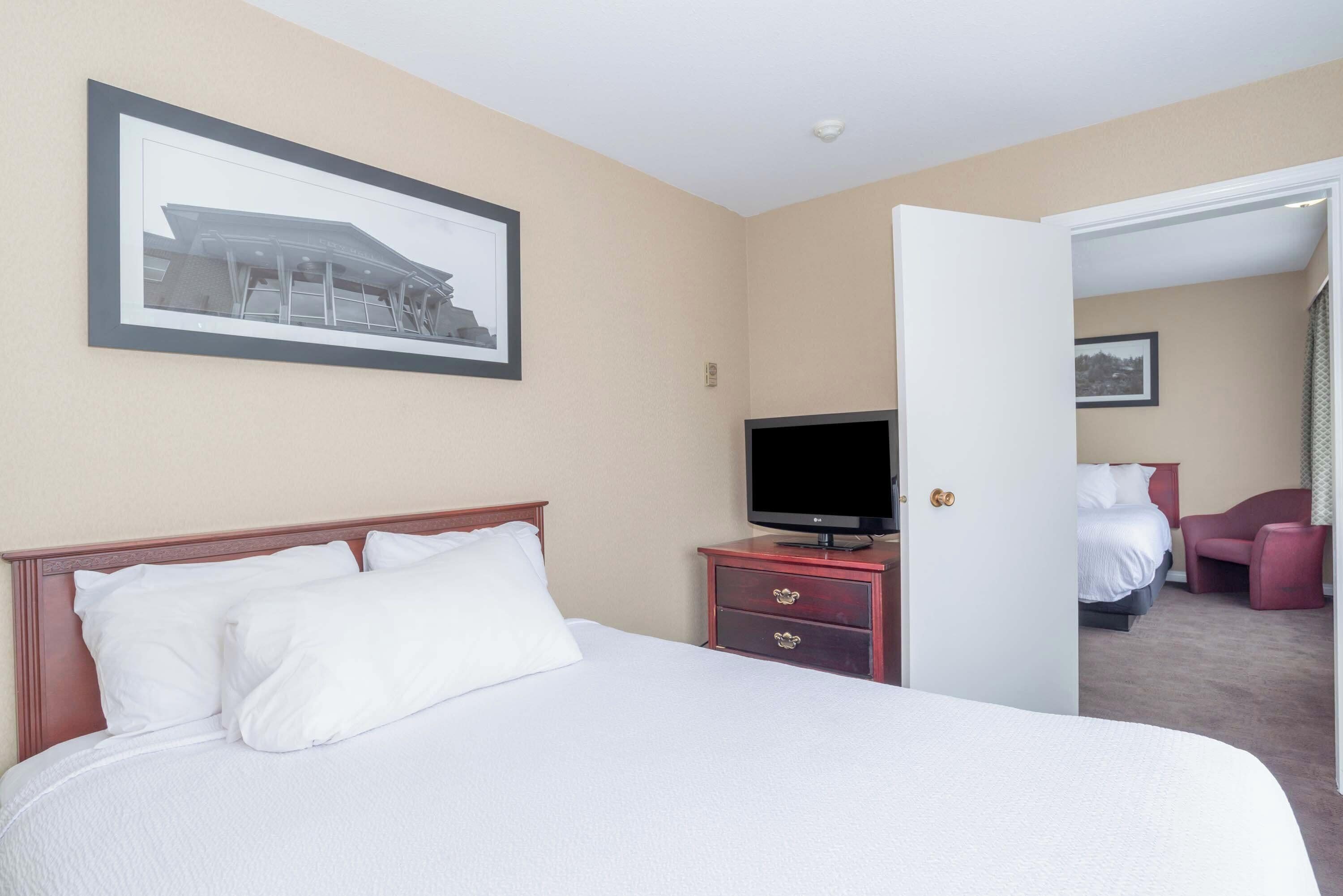 A hotel room with a bed, a television on a dresser, a framed picture on the wall, and a door leading to another room.