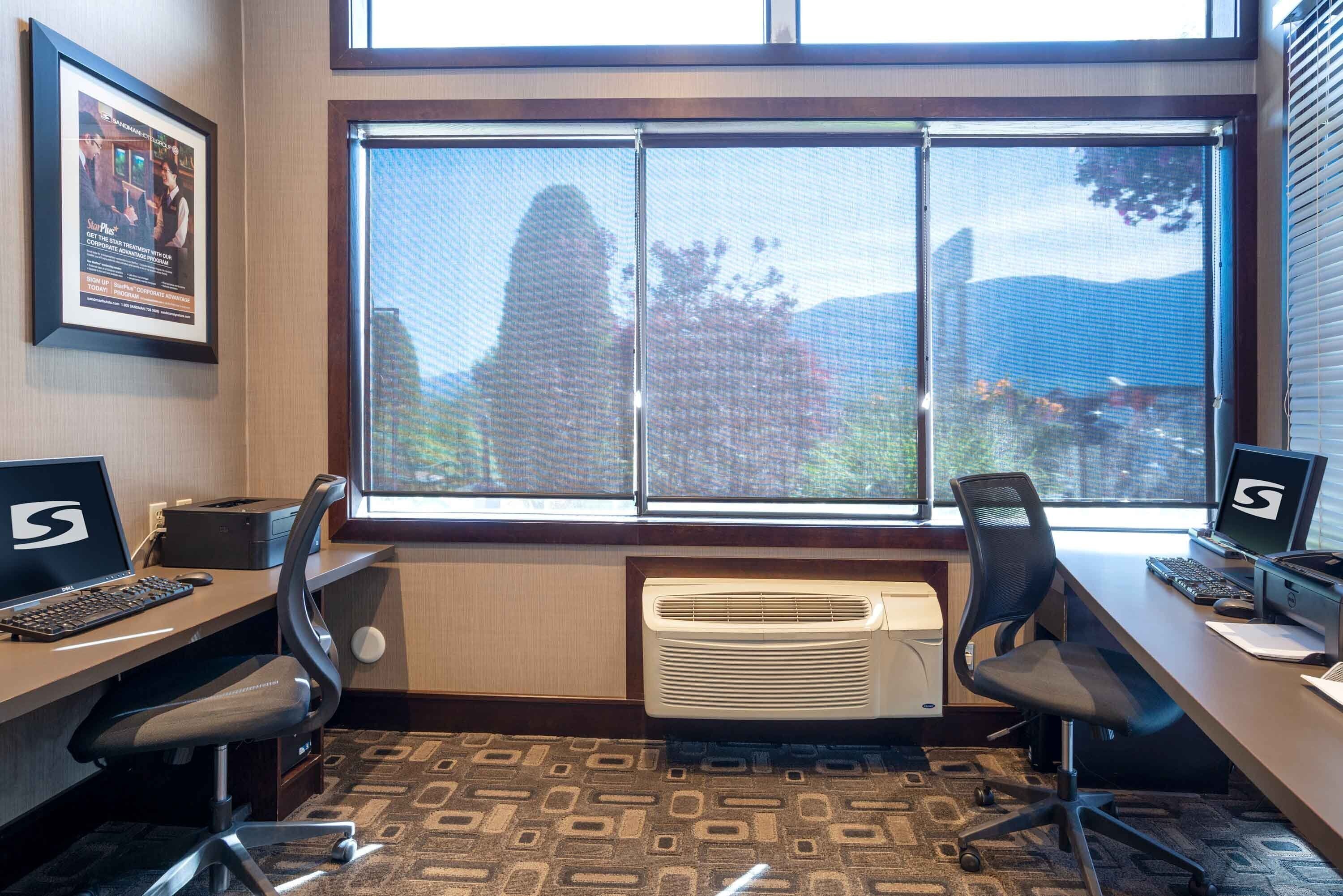 Habitación de hotel con un ventanal grande, dos escritorios con computadoras, un calentador vertical y una alfombra estampada.
