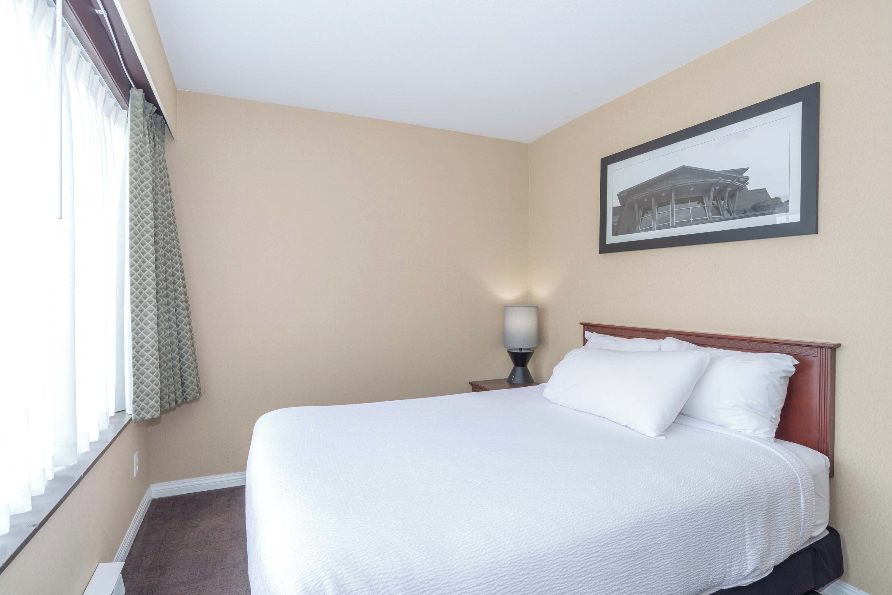 A hotel room with a bed, a nightstand with a lamp, a framed picture on the wall, and a window with curtains.