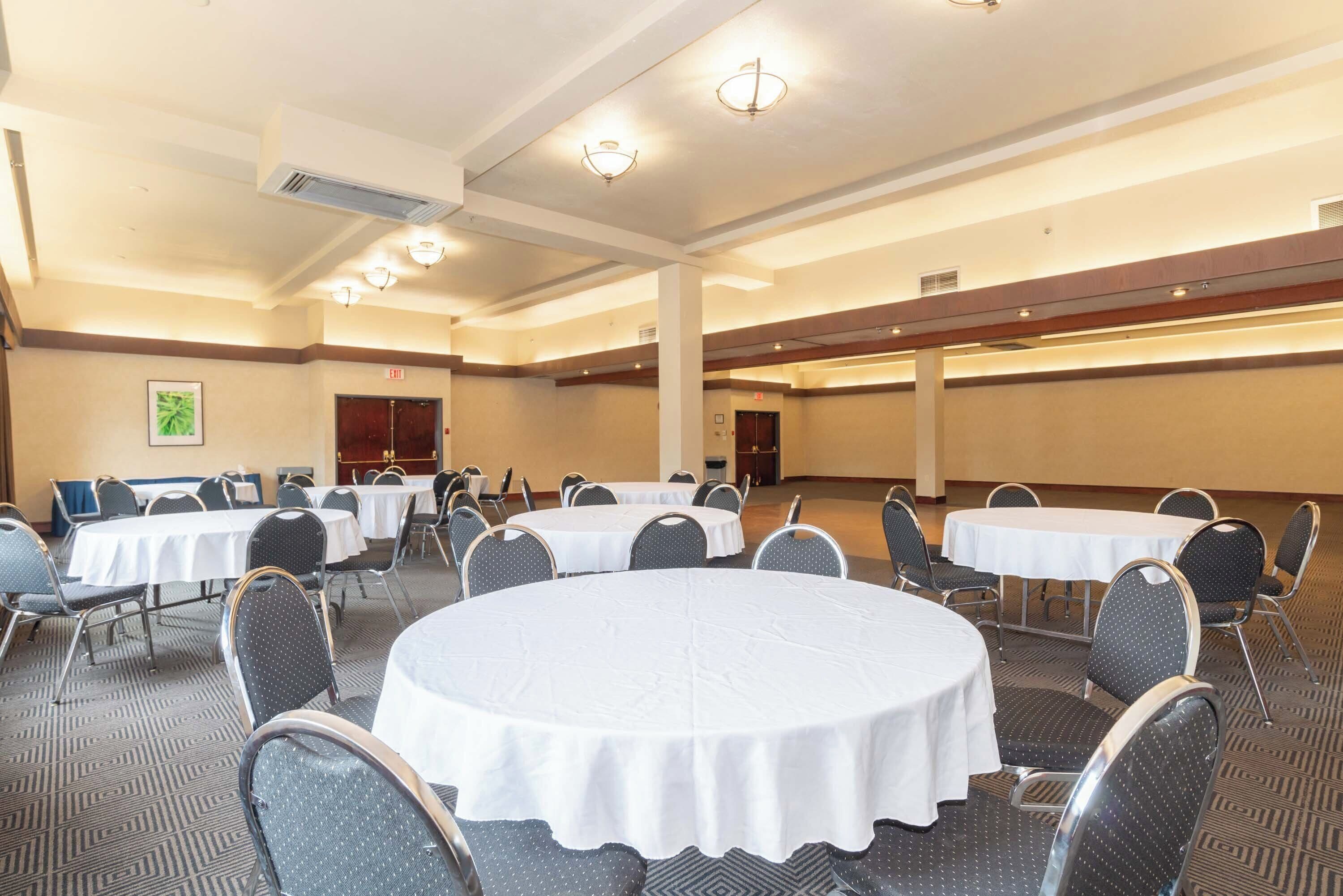 A conference room with multiple round tables, each surrounded by chairs. There are ceiling lights and an air conditioning unit visible.