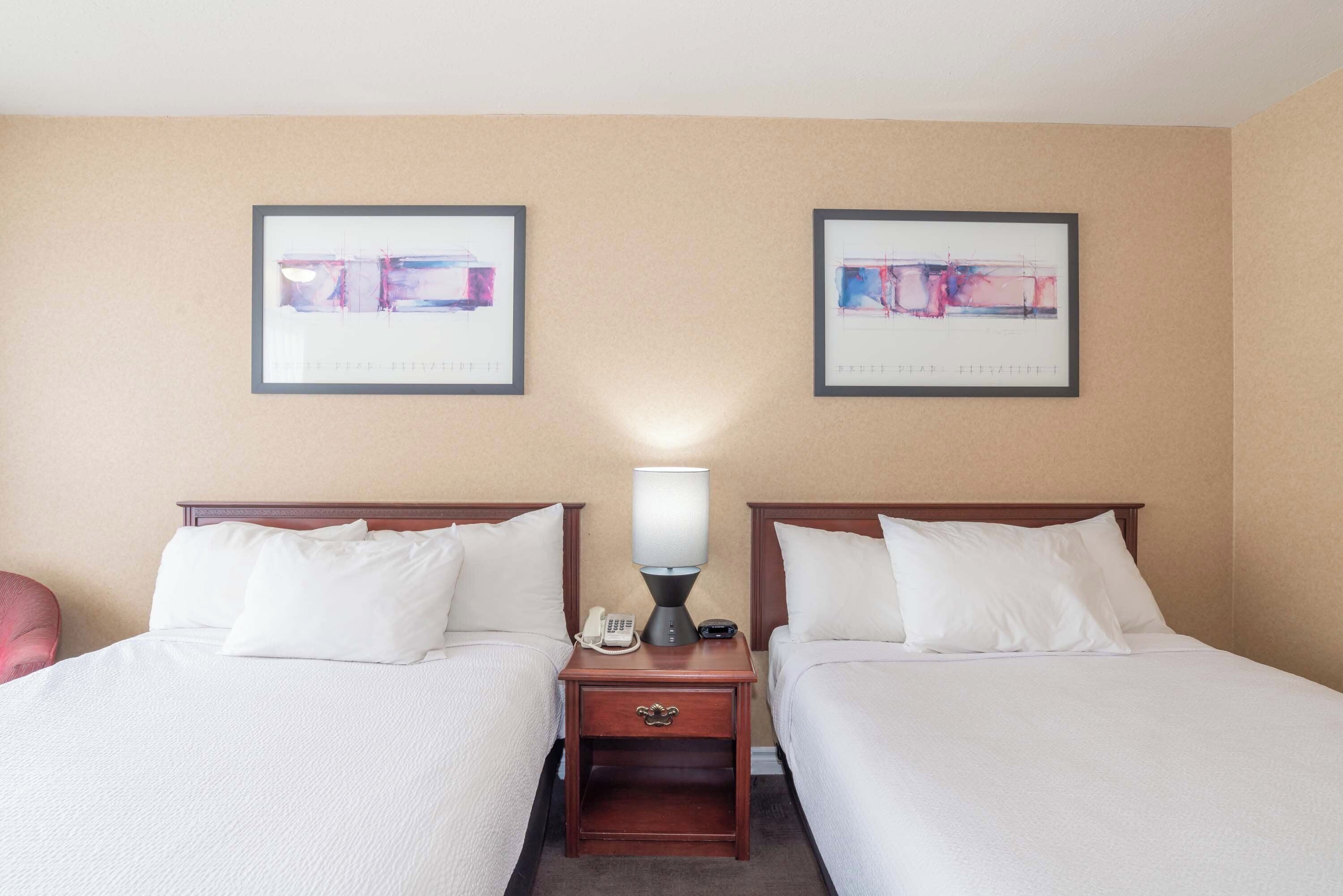 A hotel room with two beds, a nightstand with a lamp, and two framed abstract paintings on the wall.