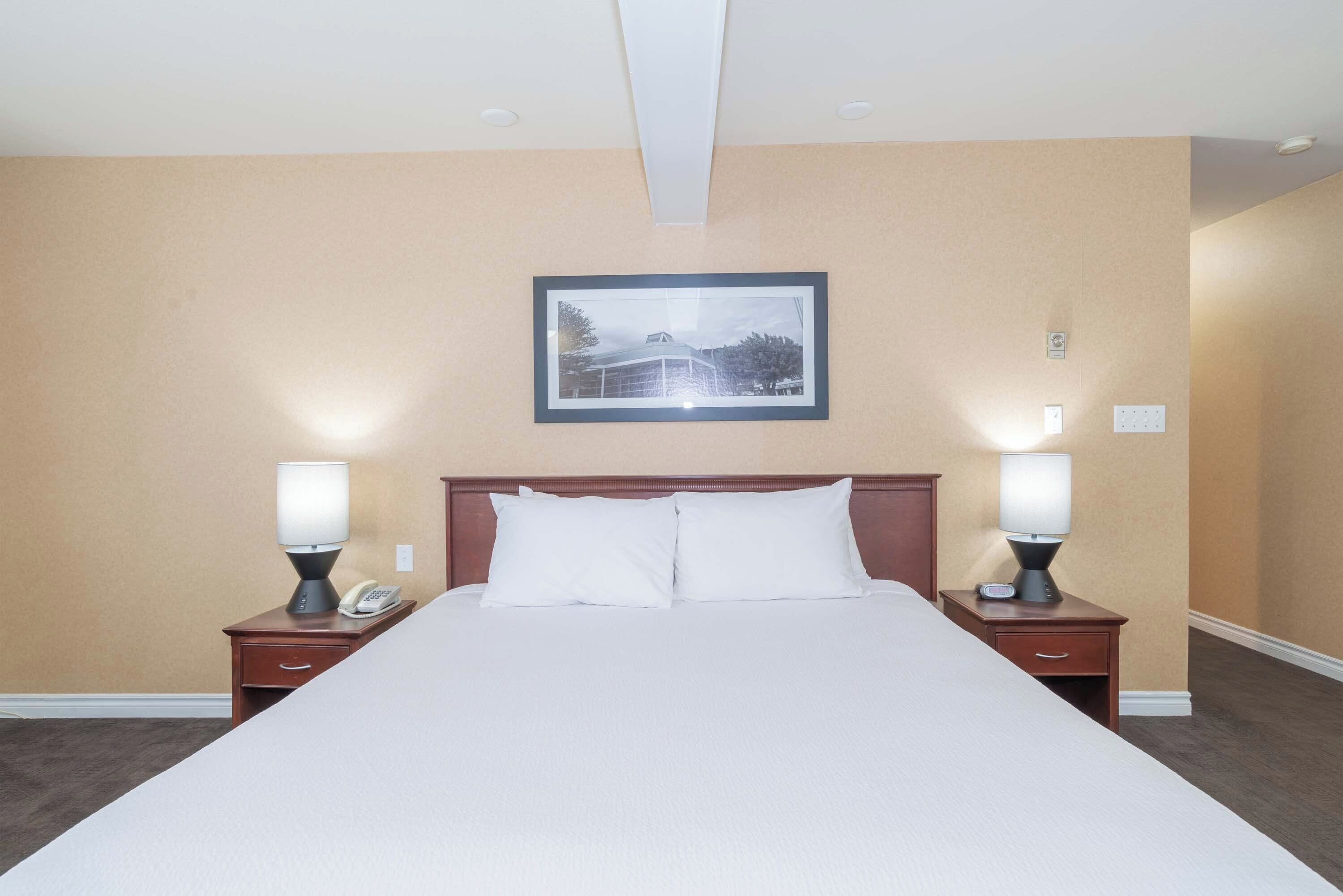Una habitación de hotel con una cama grande, dos mesitas de noche con lámparas, un cuadro enmarcado en la pared y una pared beige lisa.