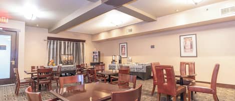 A hotel dining area with multiple tables and chairs, a patterned carpet, and ceiling beams.